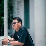 An Indonesian man in glasses daydreaming on a Jakarta balcony, captured in natural daylight.